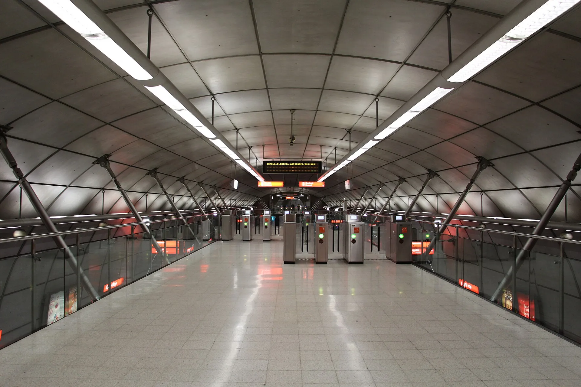 Un andén de metro con bóveda metálica, tubos de luz y torniquetes al fondo, en blanco y negro con un tono verde.